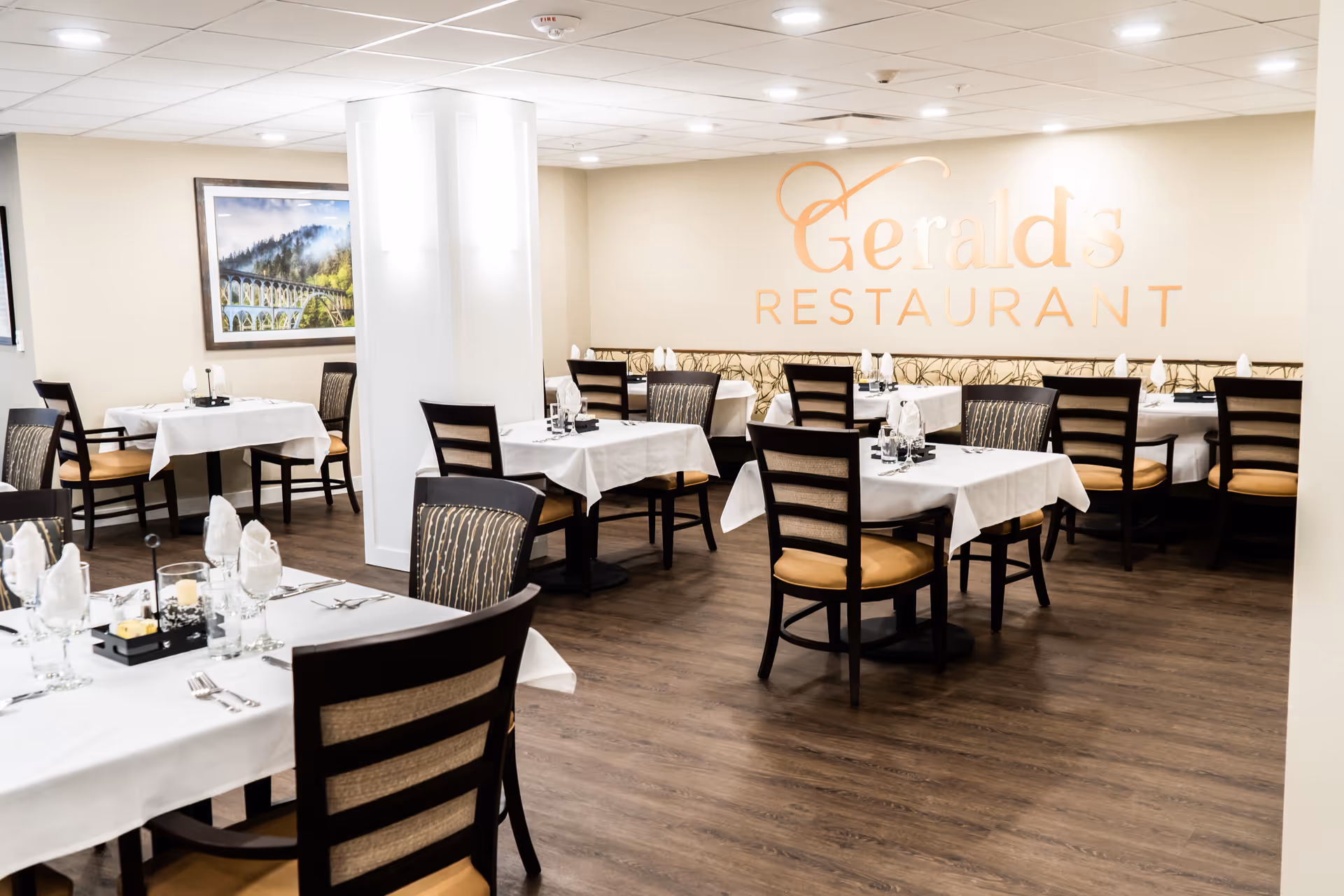 Interior view of Gerald's Restaurant with neatly arranged tables covered with white tablecloths, set with glasses, napkins, and cutlery. The room has wooden flooring, beige walls, and a framed picture of a scenic bridge and forest. The restaurant name is displayed on the wall in large letters.