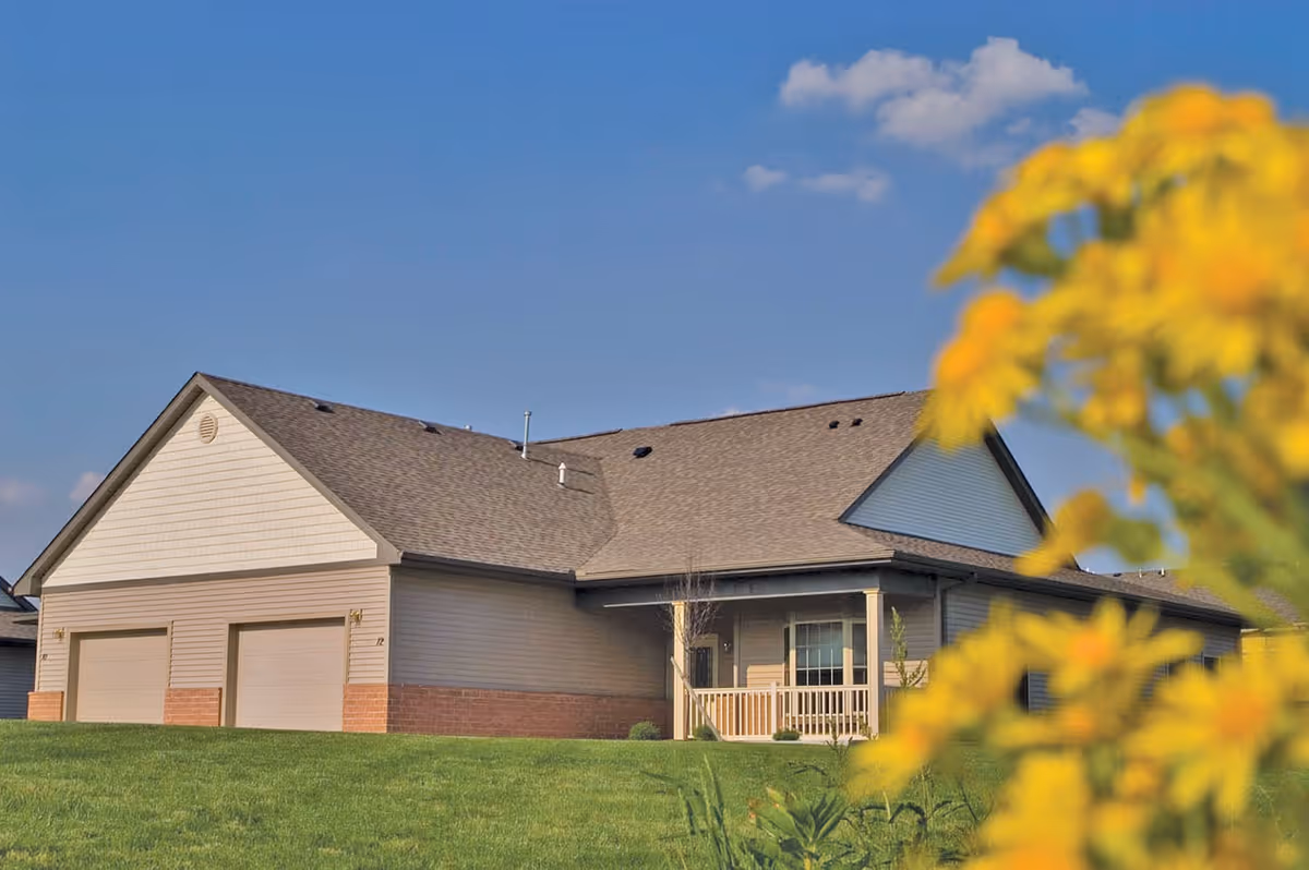 Exterior view of a single-story residential building with a two-car garage, beige siding, and a front porch. The building is surrounded by a green lawn and yellow flowers in the foreground under a clear blue sky.