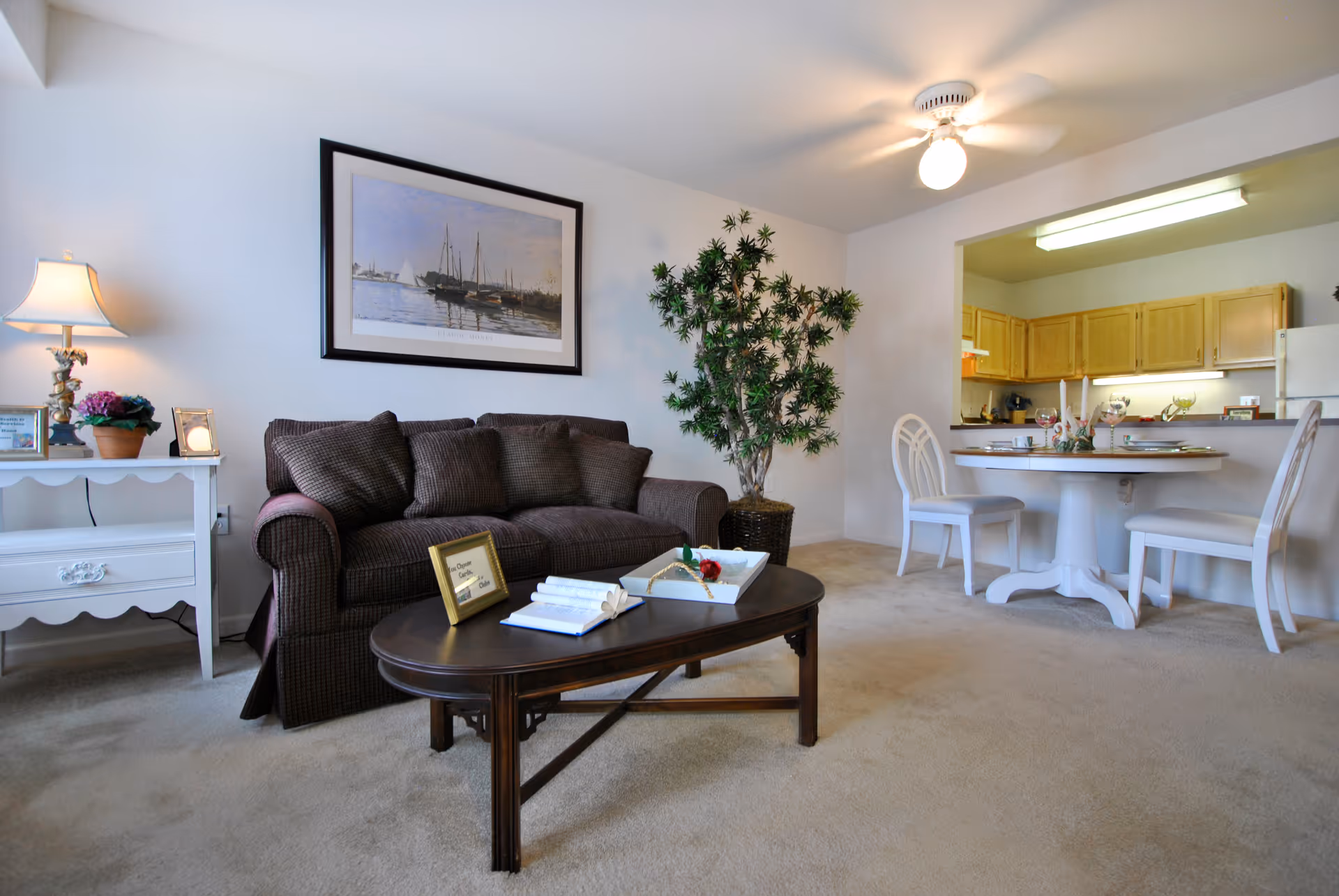 Open-plan living area with a brown upholstered sofa, wooden coffee table, side table with lamp, potted plant, and a small dining table and chairs by the kitchen.