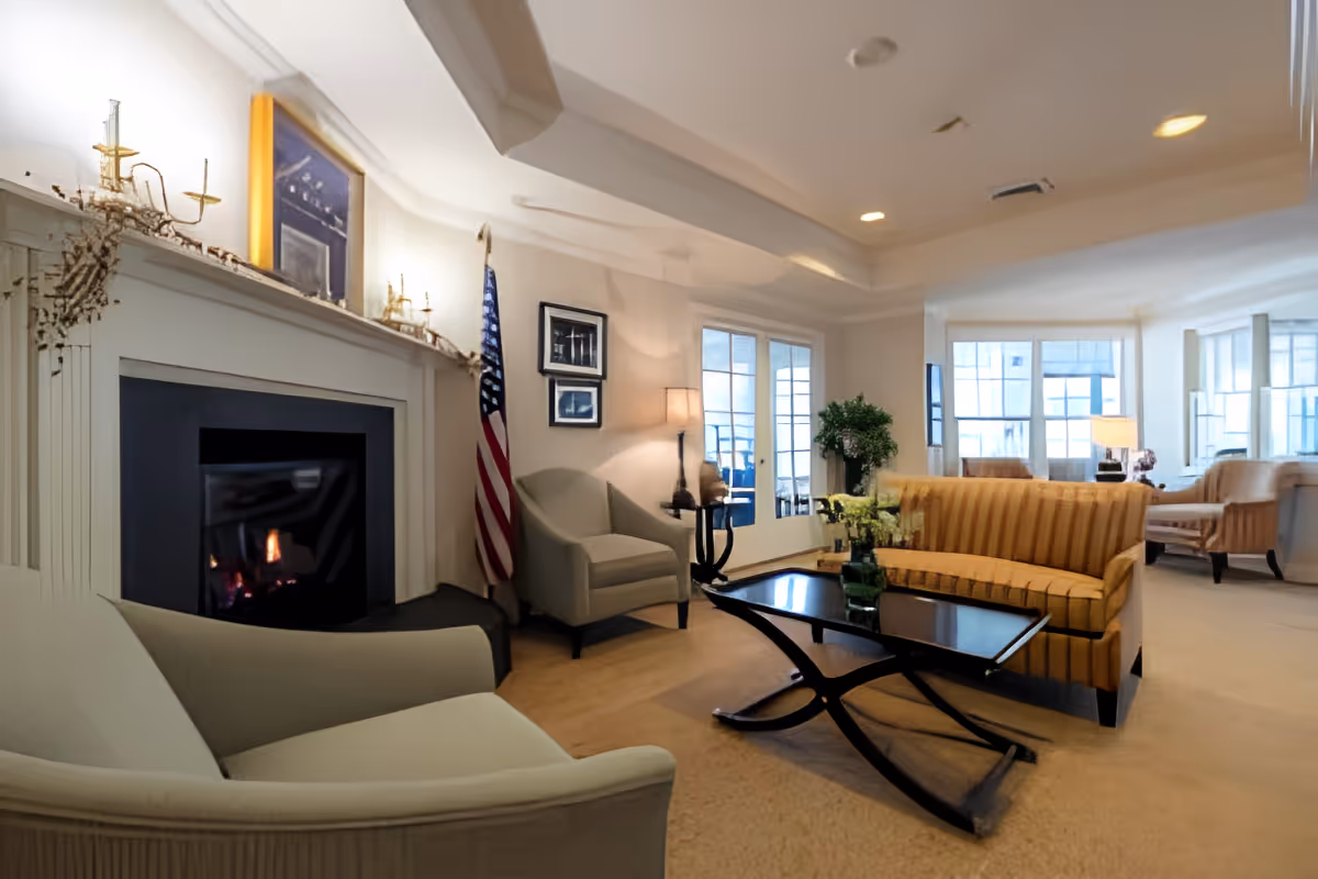 Sunlit communal living room with a fireplace, striped sofa, armchairs, coffee table, and an American flag.