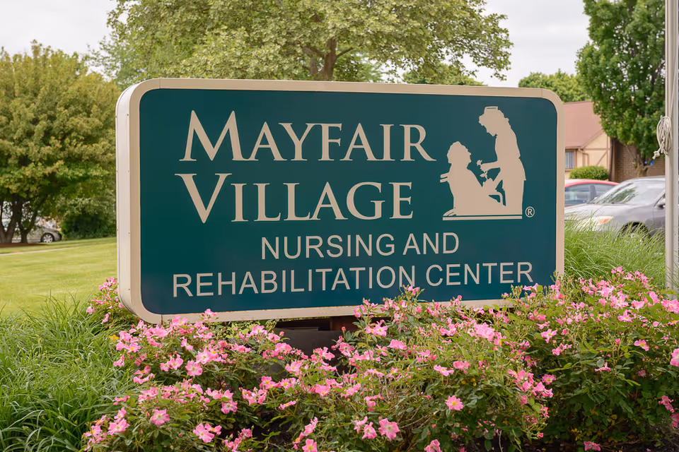 A green sign with white text that reads 'Mayfair Village Nursing and Rehabilitation Center' surrounded by pink flowers and greenery, with trees and parked cars in the background.