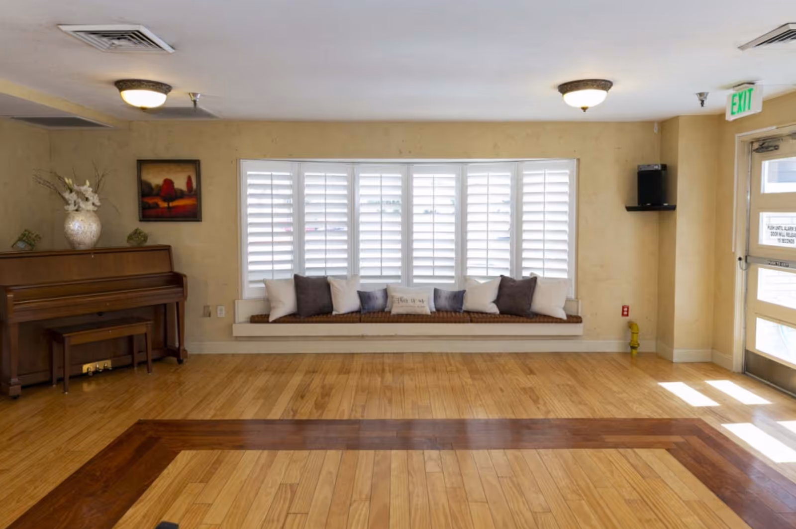 A bright room with wooden flooring featuring a built-in window bench with multiple cushions in front of white plantation shutters. To the left, there is a wooden piano with a matching bench and a large vase with flowers on top. A framed painting hangs on the wall above the piano. On the right side, there is a glass door with an exit sign above it, allowing natural light to enter the room.