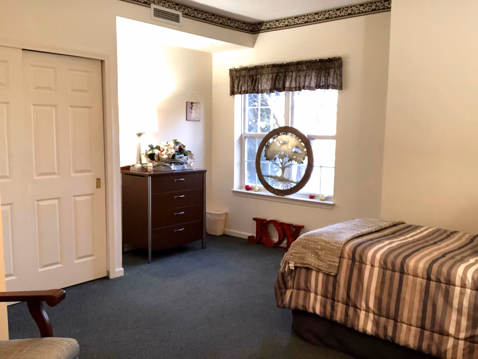 A cozy bedroom with a single bed covered in a striped comforter and a folded blanket at the foot. There is a window with a valance curtain, a decorative tree artwork hanging in front of the window, and the word 'JOY' displayed on the floor beneath the window. A wooden dresser with a lamp and a flower arrangement sits against the wall next to a sliding closet door. The room has carpeted flooring and light-colored walls.