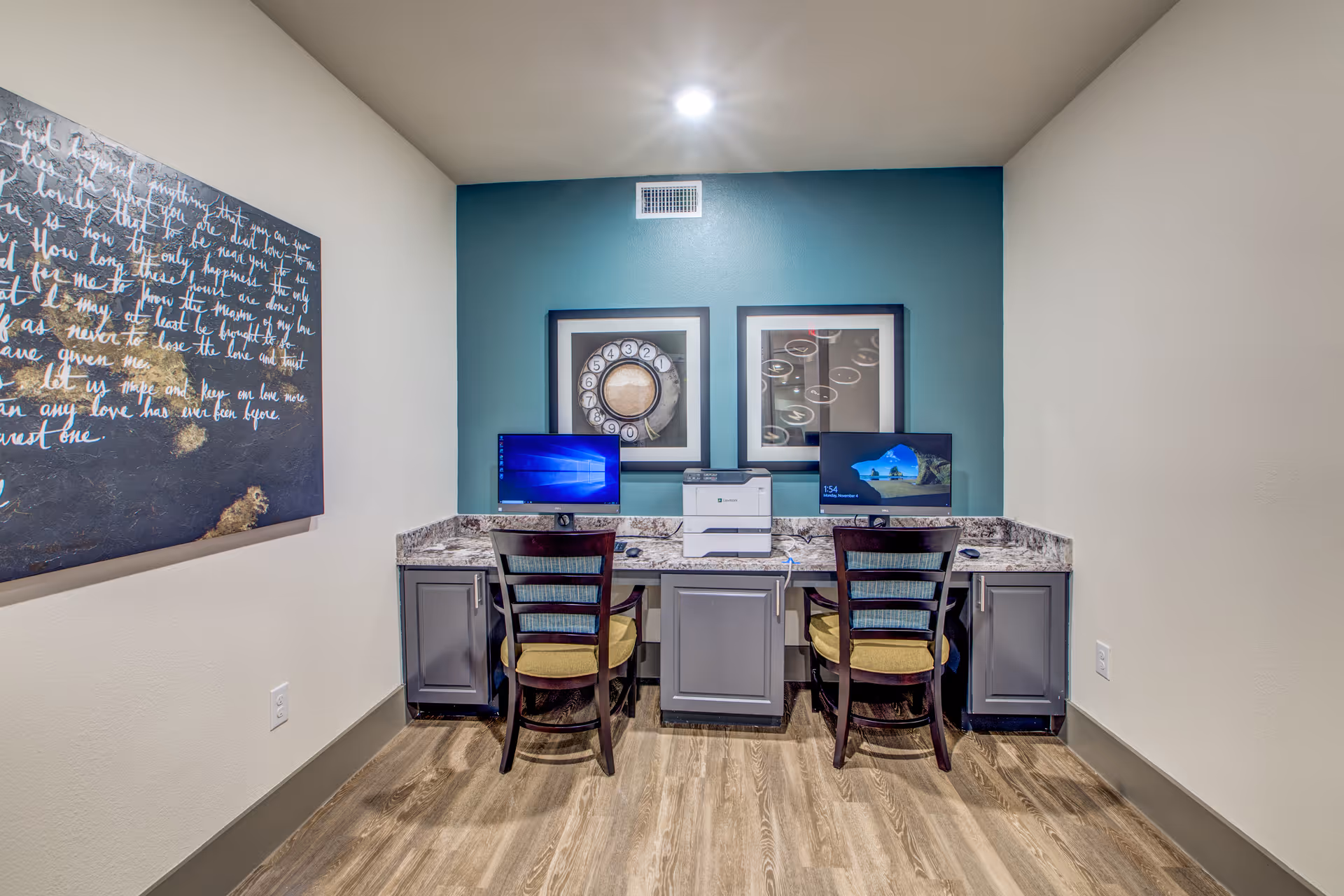 A small office area with two computer workstations and a printer on a granite countertop. Two wooden chairs with cushions are placed in front of the desks. The wall behind the desks is painted teal and decorated with two framed pictures. The floor is wood, and there is a large piece of wall art with cursive writing on the left wall.