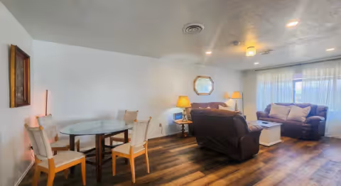 Bright living room with wood-look flooring, two sofas, a coffee table, lamps, and a small round dining table with four chairs near a curtained window.