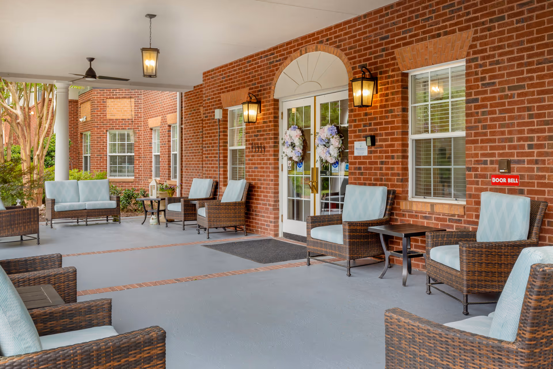 Covered brick entrance patio with wicker chairs, side tables, and double doors decorated with floral wreaths.