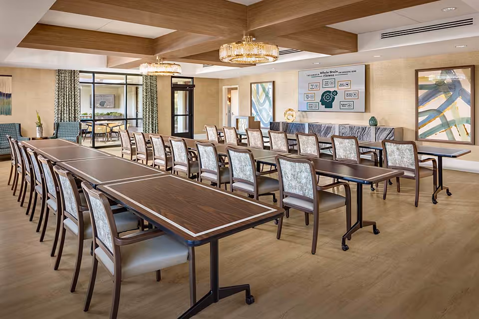 A spacious meeting or activity room with multiple rows of wooden tables and cushioned chairs arranged neatly. The room features modern chandeliers, large abstract paintings on the walls, and a large screen displaying information about brain fitness. There are large windows and a glass door leading to another room with additional seating.