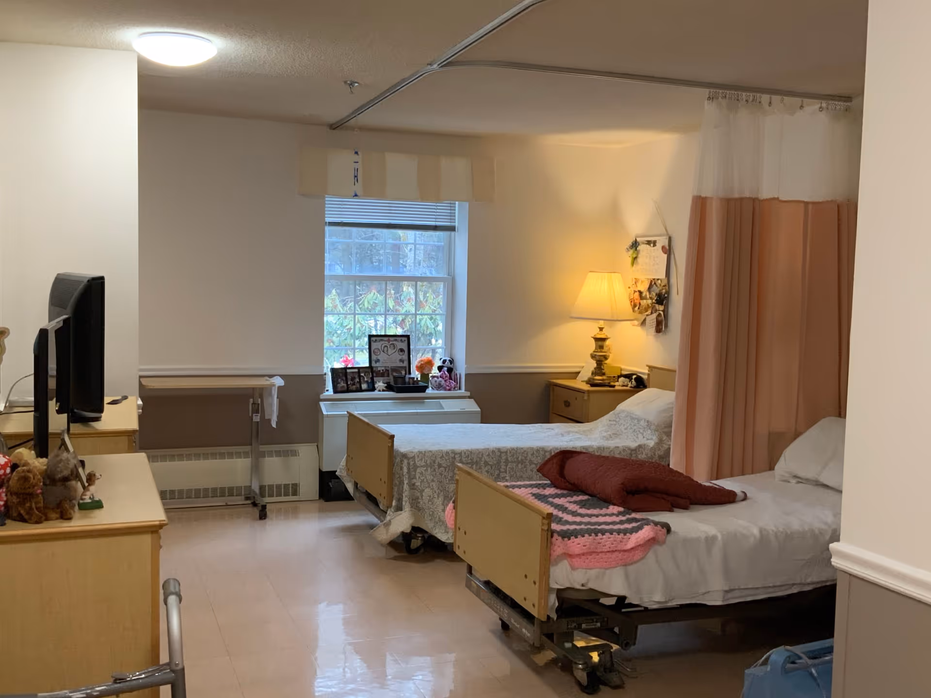 A shared bedroom in a care facility with two hospital-style beds separated by a privacy curtain. Each bed has blankets and pillows, and there is a bedside table with a lamp and personal items. A window with blinds lets in natural light, and a dresser with a TV and stuffed animals is visible.
