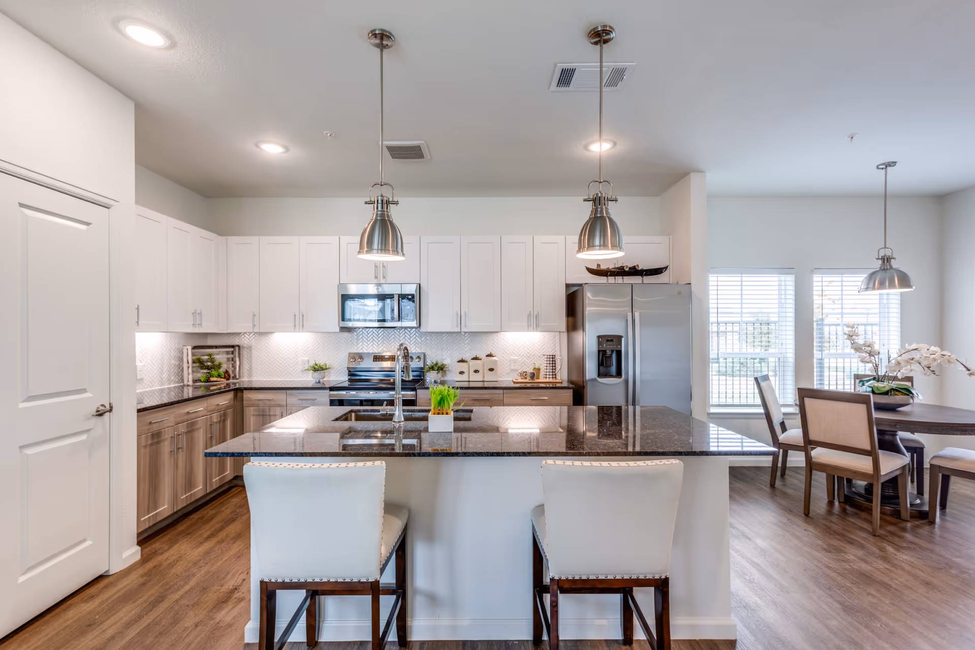 Modern kitchen with white upper cabinets and wood lower cabinets, a large island with a dark granite countertop, two white cushioned bar stools, stainless steel appliances including a refrigerator and microwave, pendant lights hanging above the island, and a dining area with a round table and chairs near windows with blinds.
