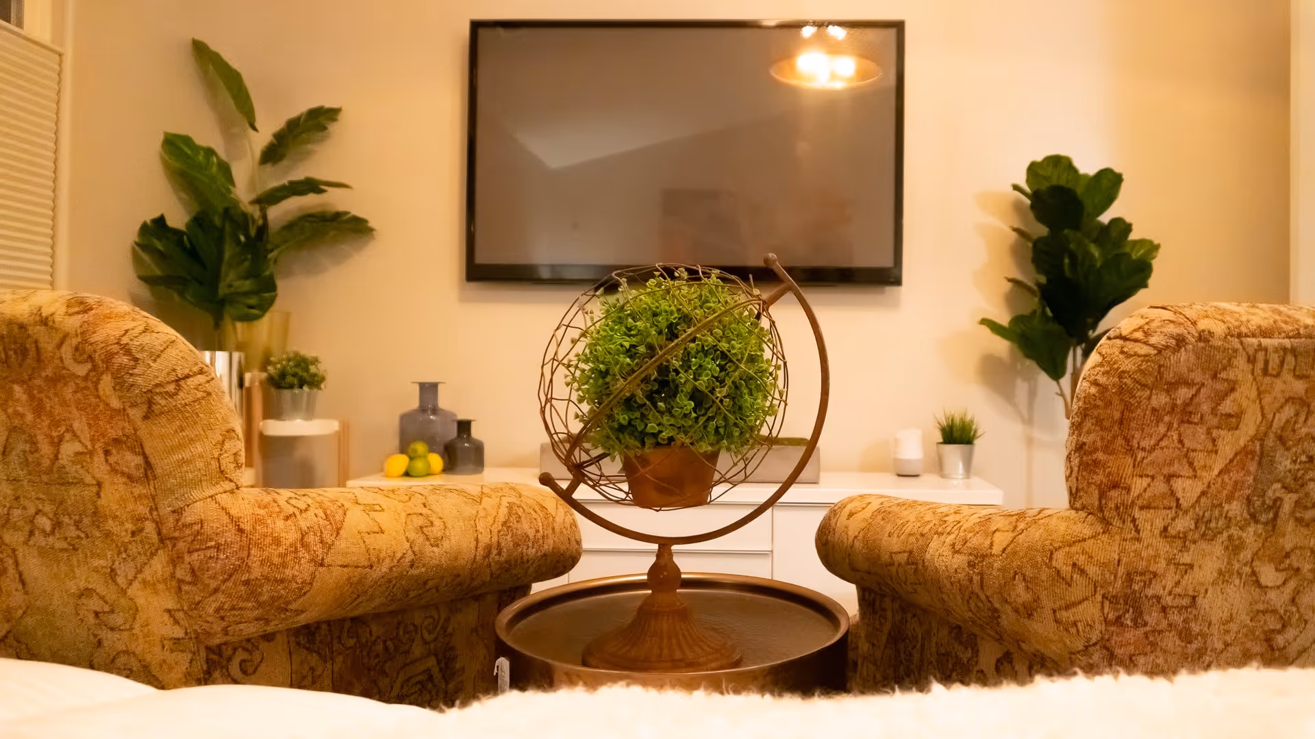 A cozy living room with two patterned armchairs facing a wall-mounted flat-screen TV. Between the chairs is a round side table with a decorative globe holding a green plant. The background features a white cabinet with decorative vases, lemons, and small plants, along with two large leafy plants on either side.
