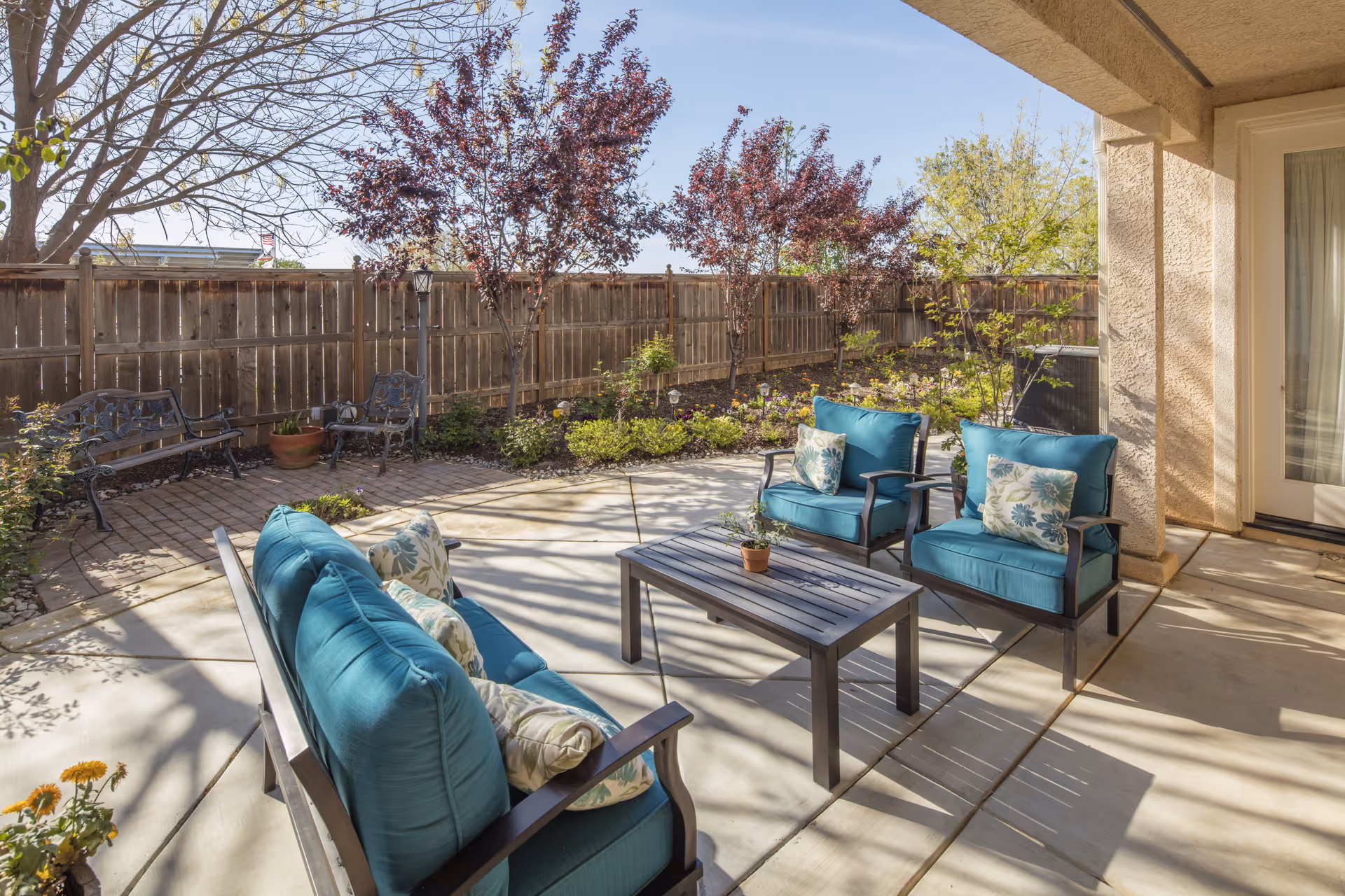 Outdoor patio area with cushioned seating including a sofa and two armchairs with blue cushions and floral pillows, a rectangular coffee table, surrounded by a garden with small trees and plants, enclosed by a wooden fence under a clear sky.