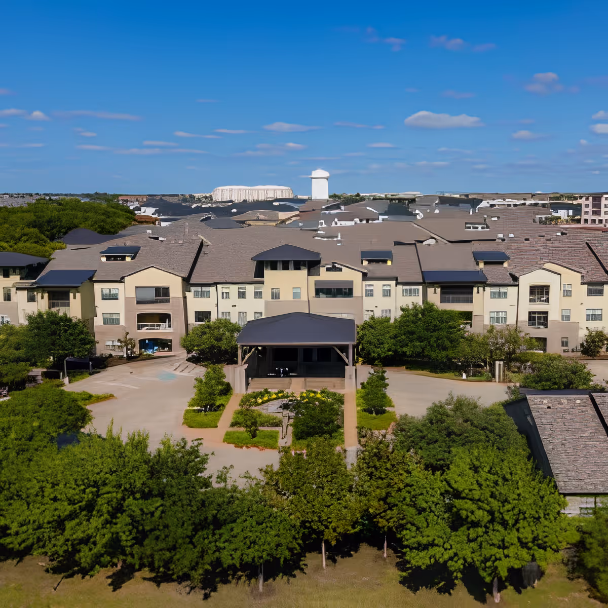 Aerial view of Discovery Village At Twin Creeks, showing a large multi-story residential building surrounded by trees and greenery under a blue sky with scattered clouds.