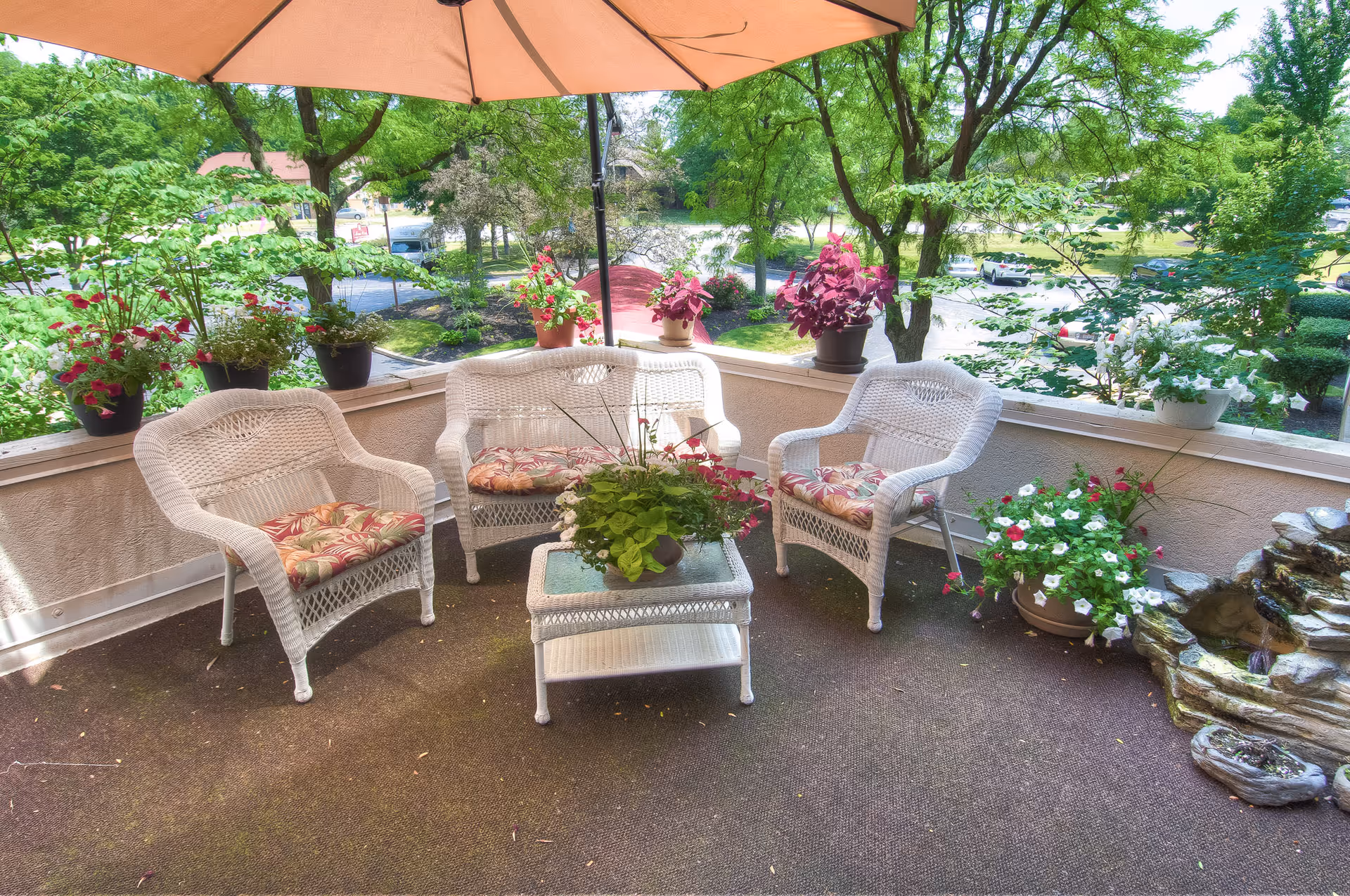 Outdoor patio area with white wicker furniture including two chairs and a loveseat with floral cushions, a glass-top wicker table with a potted plant, surrounded by various potted flowers and plants. There is a large umbrella providing shade and trees and a parking lot visible in the background.