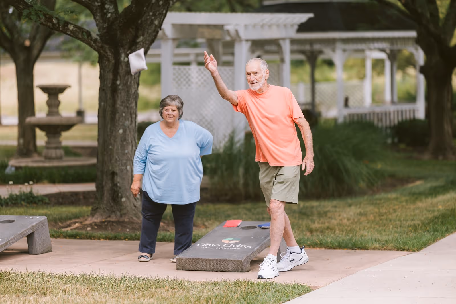 An elderly man and woman playing cornhole outdoors on a paved area with grass and trees around. The man is in mid-throw, wearing a coral t-shirt, khaki shorts, and white sneakers. The woman stands nearby watching, wearing a light blue top and dark pants. A white pergola and a stone fountain are visible in the background.