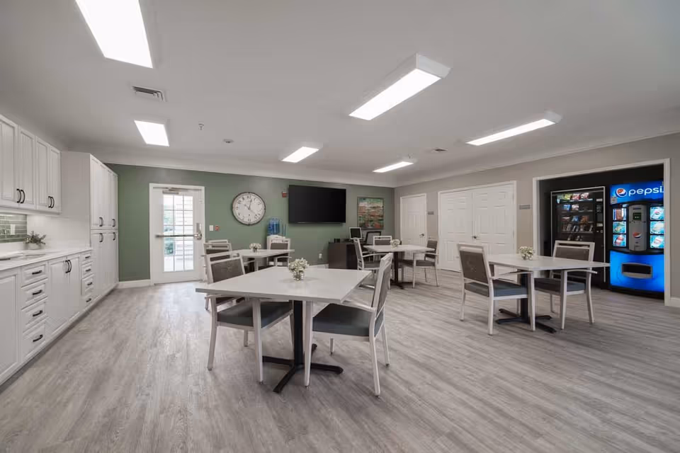 A clean and modern dining room with several square tables and chairs arranged neatly. The room has light wood flooring, white cabinetry along one wall, a large clock, a flat-screen TV mounted on a green accent wall, and a vending machine area with Pepsi branding. There is also a water dispenser and some decorative flowers on the tables.