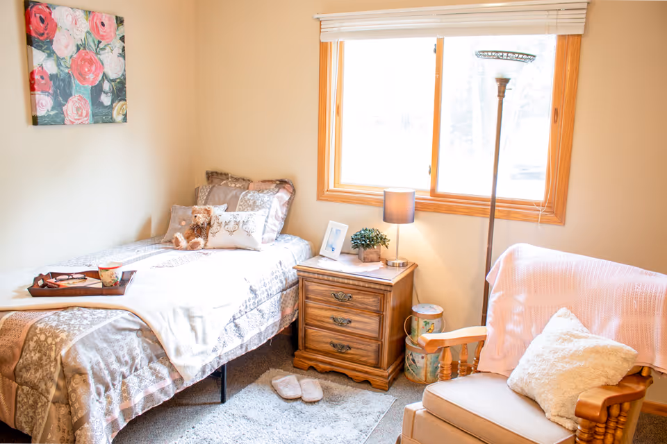 A cozy bedroom with a single bed covered in patterned bedding and a teddy bear on the pillows. A wooden nightstand beside the bed holds a small lamp, a framed photo, and a small plant. Next to the nightstand is a tall floor lamp and a comfortable wooden armchair with a pink throw blanket and a white pillow. A window with wooden trim lets in natural light, and a floral painting hangs on the wall above the bed.