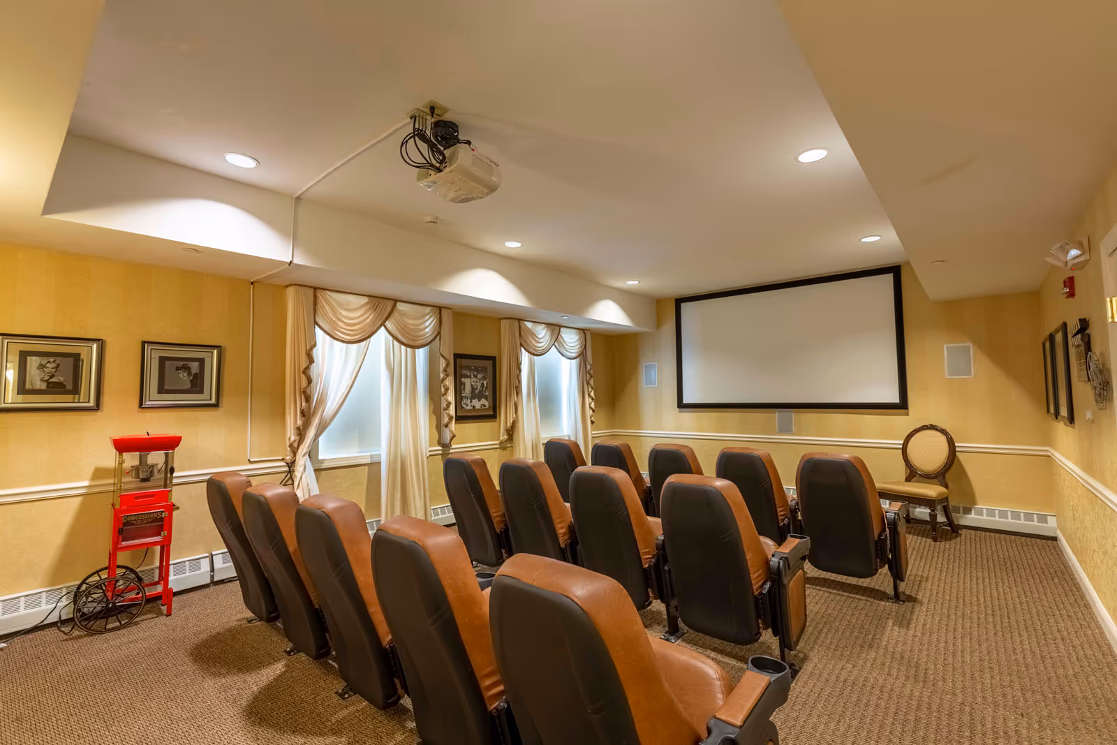 A small theater room with three rows of brown leather seats facing a large blank projection screen. The room has beige walls with white trim, carpeted floor, and two windows with cream-colored drapes. A popcorn machine is positioned against the left wall, and framed black and white pictures hang on the walls. A ceiling-mounted projector is visible above the seats.