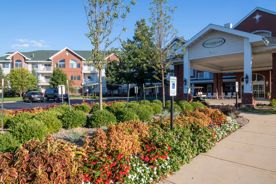Exterior view of Touchmark on West Prospect facility showing a landscaped garden with colorful flowers and bushes in front of a covered entrance with white pillars and a sign displaying the facility name and address. The building has red brick and white siding with balconies and green roofs under a clear blue sky.