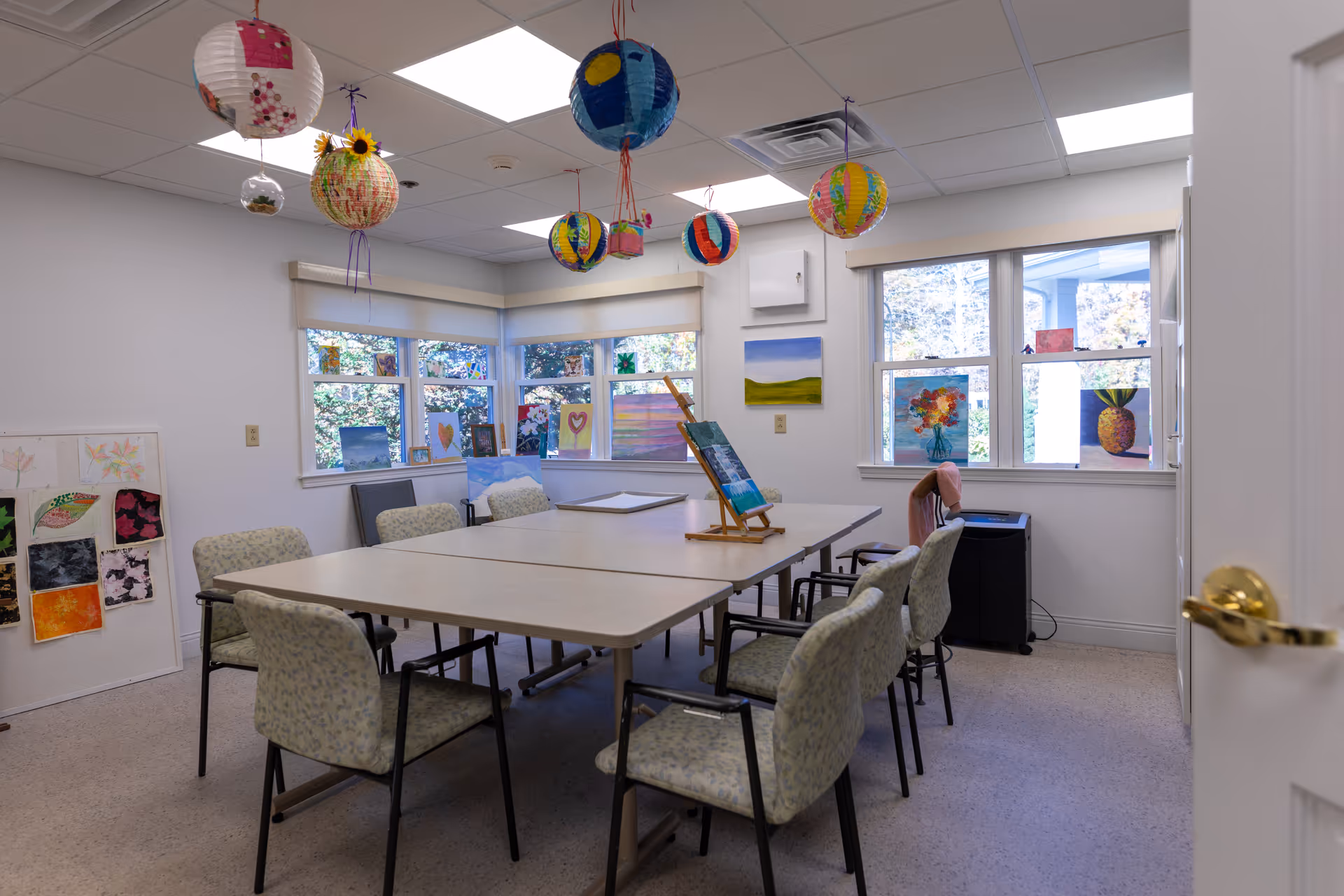 A bright art activity room with a large rectangular table surrounded by eight cushioned chairs. Colorful paper lanterns hang from the ceiling. The walls and windowsills display various paintings and artwork, including flowers and landscapes. Large windows let in natural light, and a door with a brass handle is partially open.