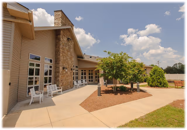Exterior view of a senior living facility building with beige siding and a stone chimney. There are several white benches along the walkway, a small landscaped area with trees and shrubs, and a clear blue sky with some clouds.