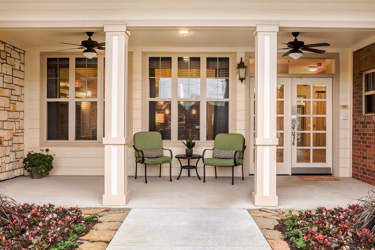 Covered entrance porch with two green chairs and a small table flanked by columns and plants.