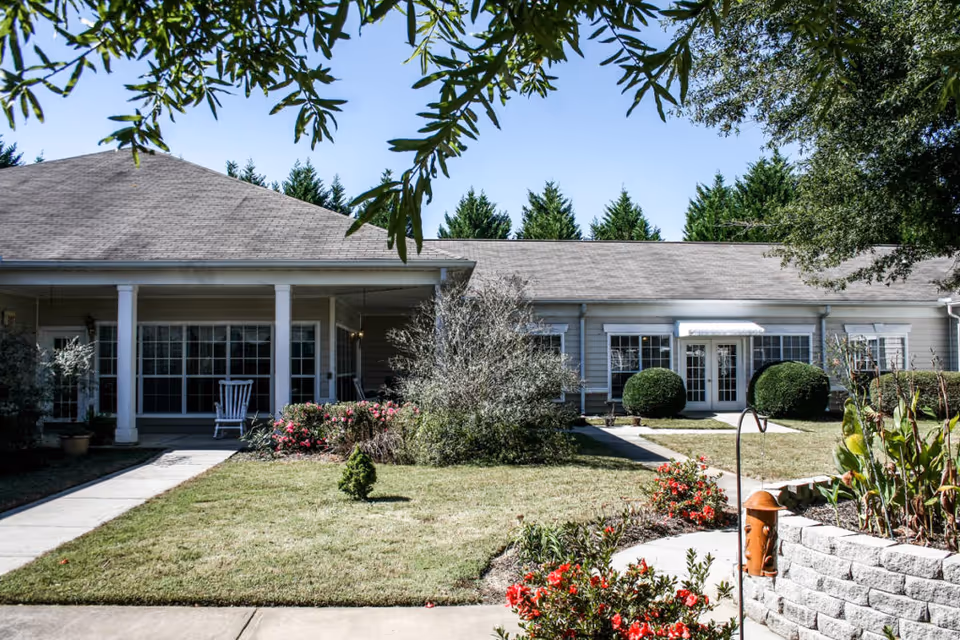 Exterior view of a single-story assisted living facility building with a covered porch, large windows, manicured lawn, bushes, and flowering plants under a clear blue sky.