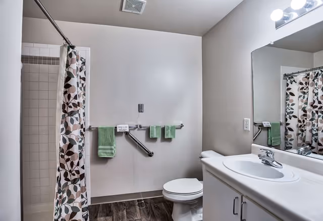 A clean bathroom with a white toilet, a white sink with a countertop, a large mirror above the sink, and a shower area with a floral-patterned shower curtain. There are green towels hanging on a metal grab bar attached to the wall, and the floor has dark wood-like tiles.