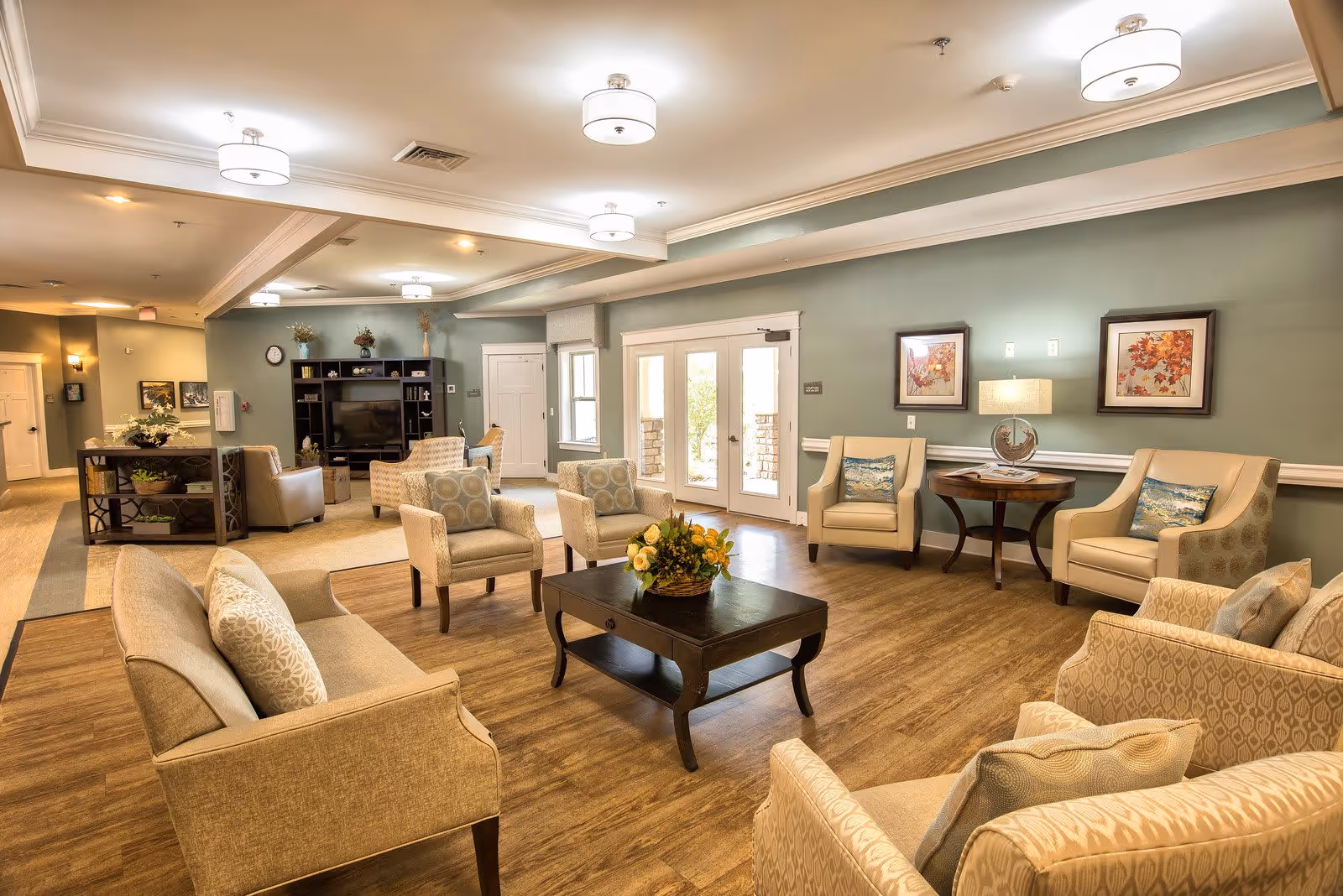 A spacious and well-lit senior living facility common area with comfortable beige armchairs and sofas arranged around a dark wooden coffee table with a floral centerpiece. The room features wood flooring, soft green walls adorned with framed artwork, a side table with a lamp, and a large entertainment center with a TV. Double glass doors lead outside, allowing natural light to fill the room.