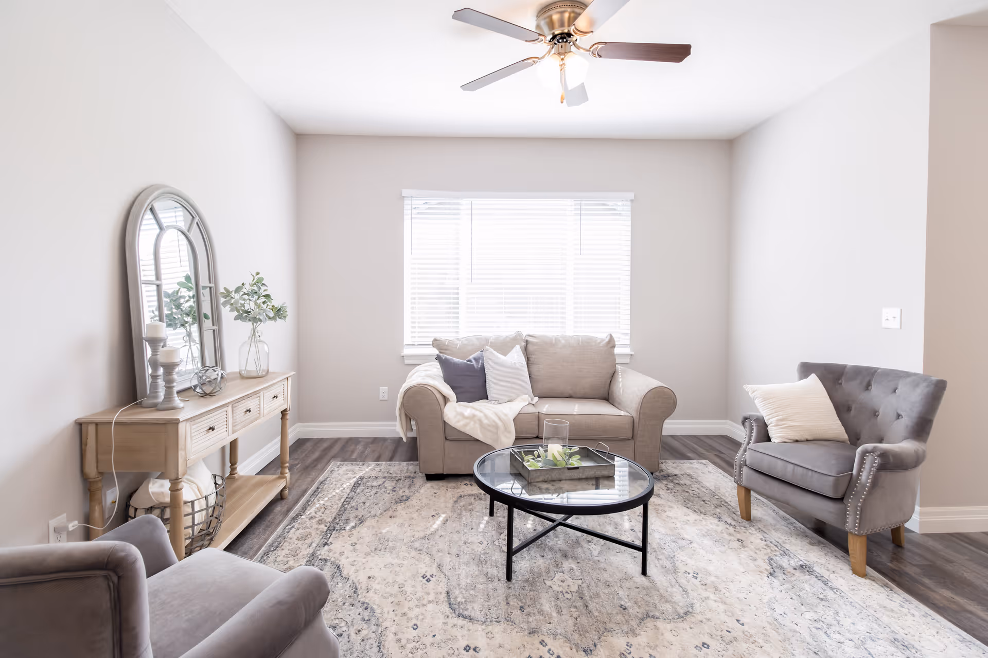 Bright neutral living room with a beige sofa, two gray armchairs, a round glass coffee table, and a console table with a mirror.