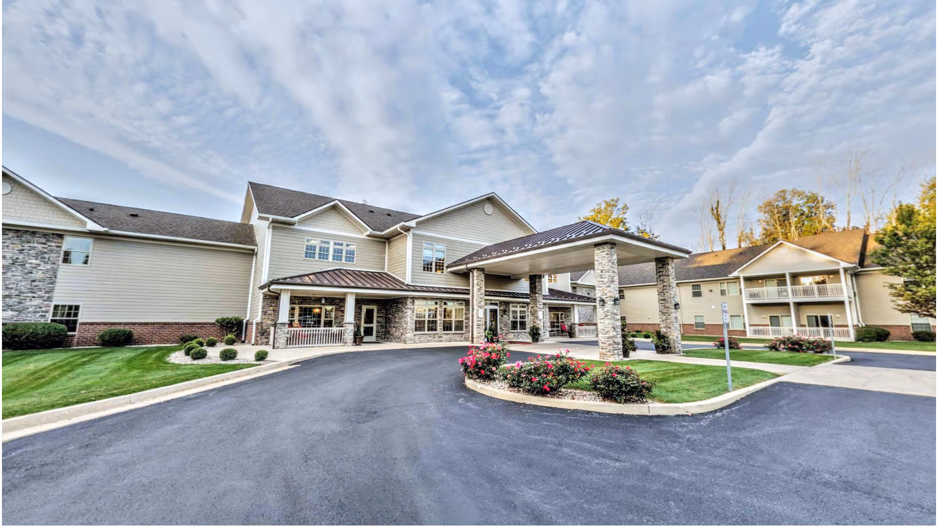 Exterior view of Primrose Retirement Community of Kokomo showing a large building with beige siding and stone accents, a covered entrance supported by stone pillars, landscaped greenery, and a paved driveway under a partly cloudy sky.