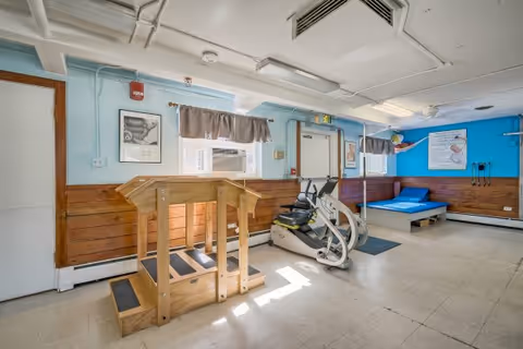 A rehabilitation or physical therapy room with light blue walls and wood paneling. The room contains a wooden step exercise platform, an exercise bike, a blue padded therapy table, and various therapy equipment mounted on the walls. There are two windows with beige curtains and fluorescent ceiling lights.