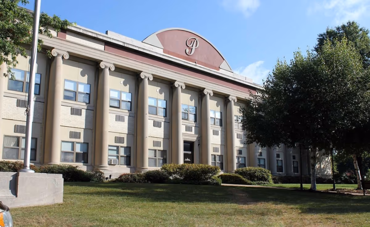 Exterior view of a large, classical-style building with tall columns and multiple windows. The building has a decorative arch at the top center with a stylized letter 'P'. There is a grassy lawn and some trees in front of the building under a clear blue sky.