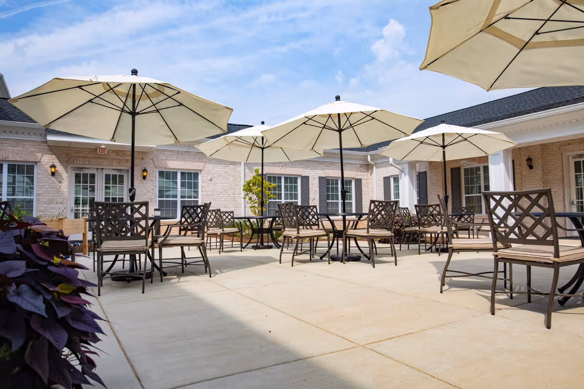 Outdoor patio area with multiple tables and chairs under large beige umbrellas, surrounded by a brick building with windows and doors under a partly cloudy sky.