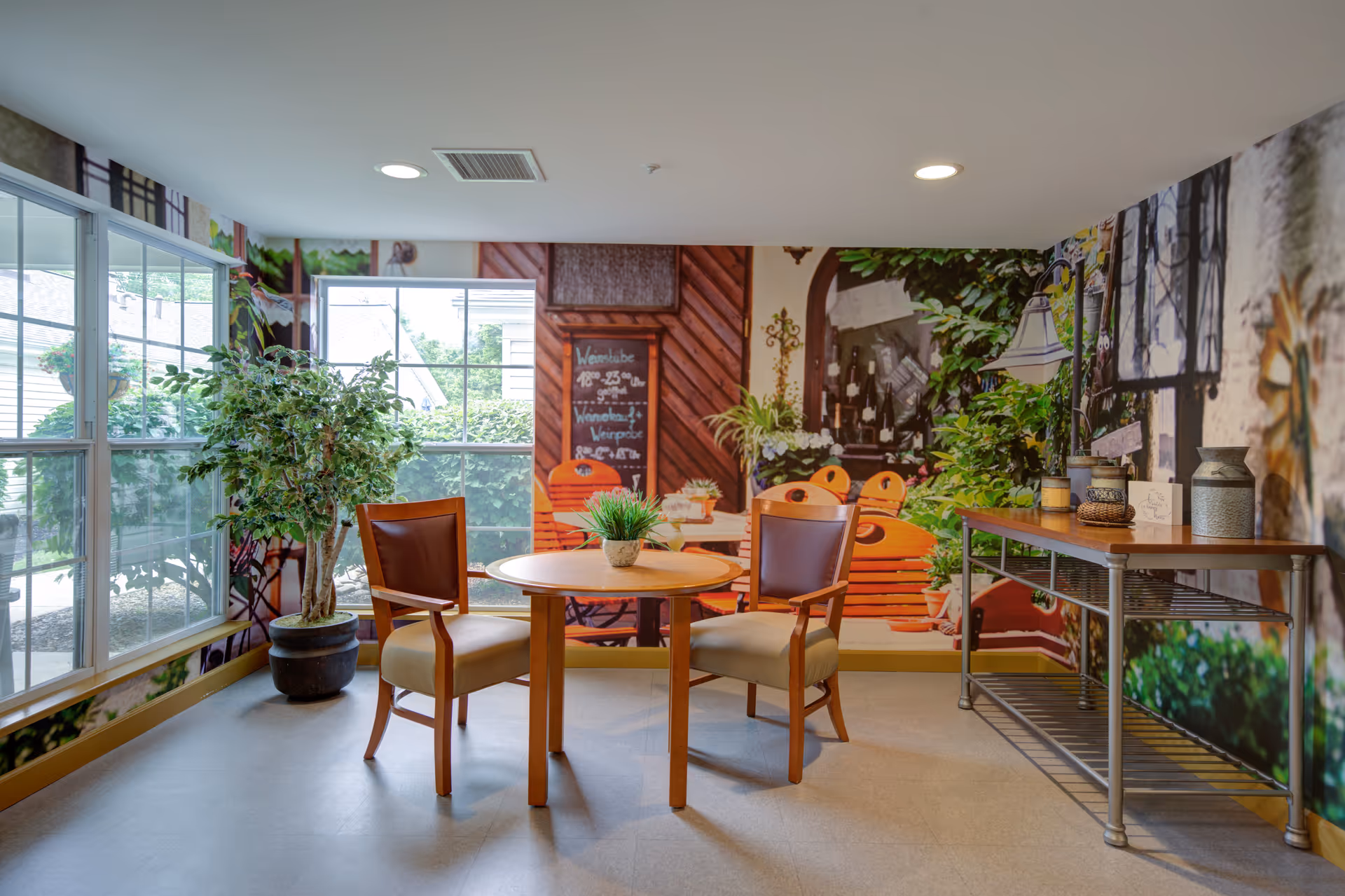A small seating area with a round wooden table and two wooden chairs with cushioned seats and backs. There is a potted plant on the table and a larger potted plant near the window. The walls have a large mural depicting an outdoor garden scene with plants, a wooden bench, and a chalkboard. A metal console table with decorative items is positioned against the right wall. Large windows allow natural light to fill the room.