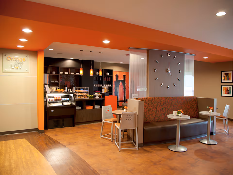 Interior view of a modern cafe area named Orange Cafe with orange accents. The space features a seating area with a cushioned bench and small round tables, a large wall clock, pendant lights, and a counter displaying pastries and beverages.