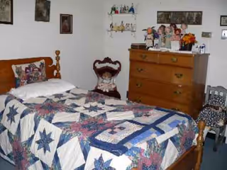 Bedroom with a quilt-covered bed, wooden dresser, and chairs against a white wall.
