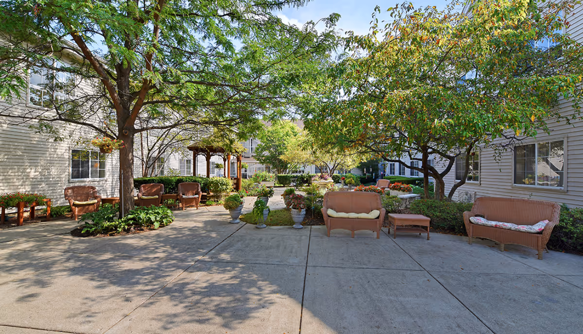 Sunny courtyard with trees, potted plants, wicker benches and chairs, and a gazebo between residential buildings.