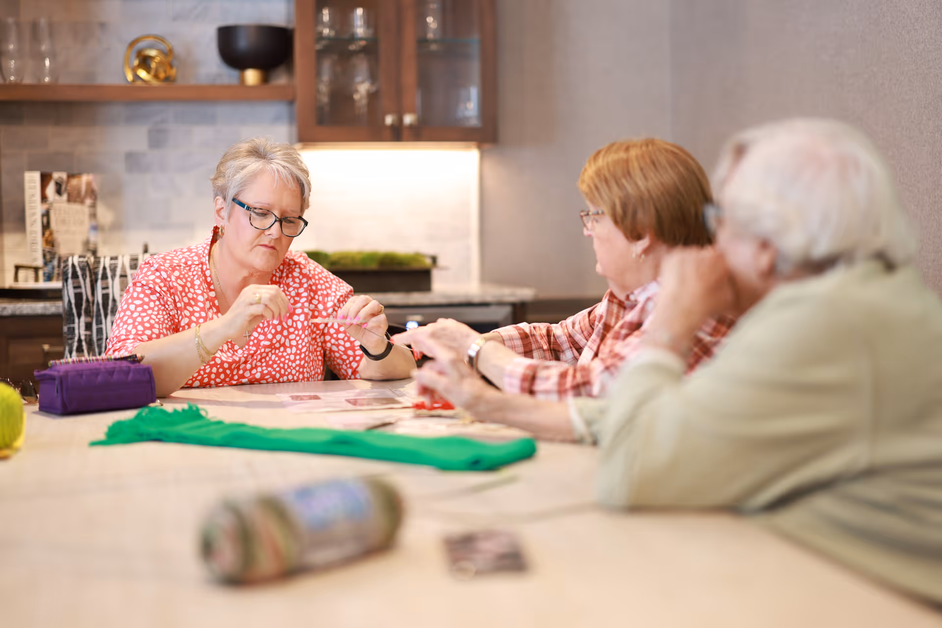 Three elderly individuals sitting around a table engaged in a crafting activity inside a room with kitchen cabinets and a countertop in the background.