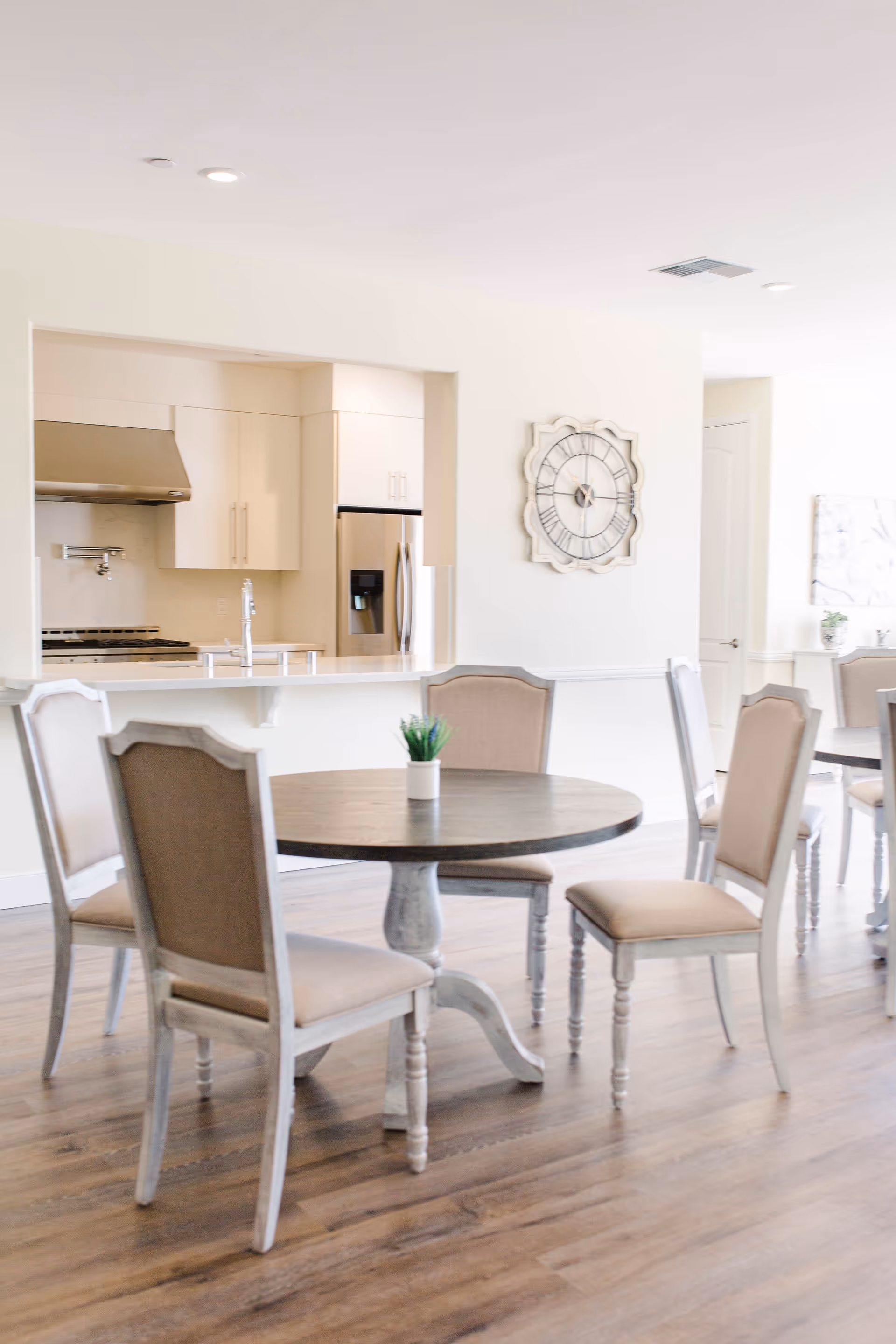A bright dining area with round wooden tables and beige upholstered chairs on a wooden floor. In the background, there is a modern kitchen with white cabinets, a stainless steel refrigerator, and a stove with a hood. A decorative wall clock is mounted on the wall between the dining area and kitchen.