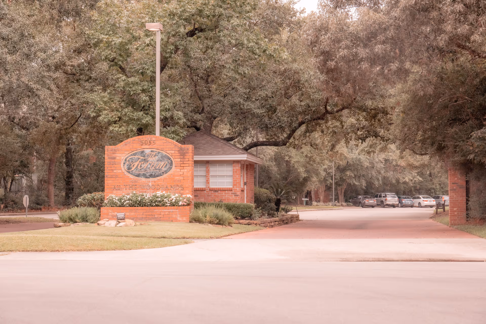 Entrance to The Forum at The Woodlands senior living facility with a brick sign and a small guardhouse surrounded by trees and greenery. Several cars are parked along the driveway.