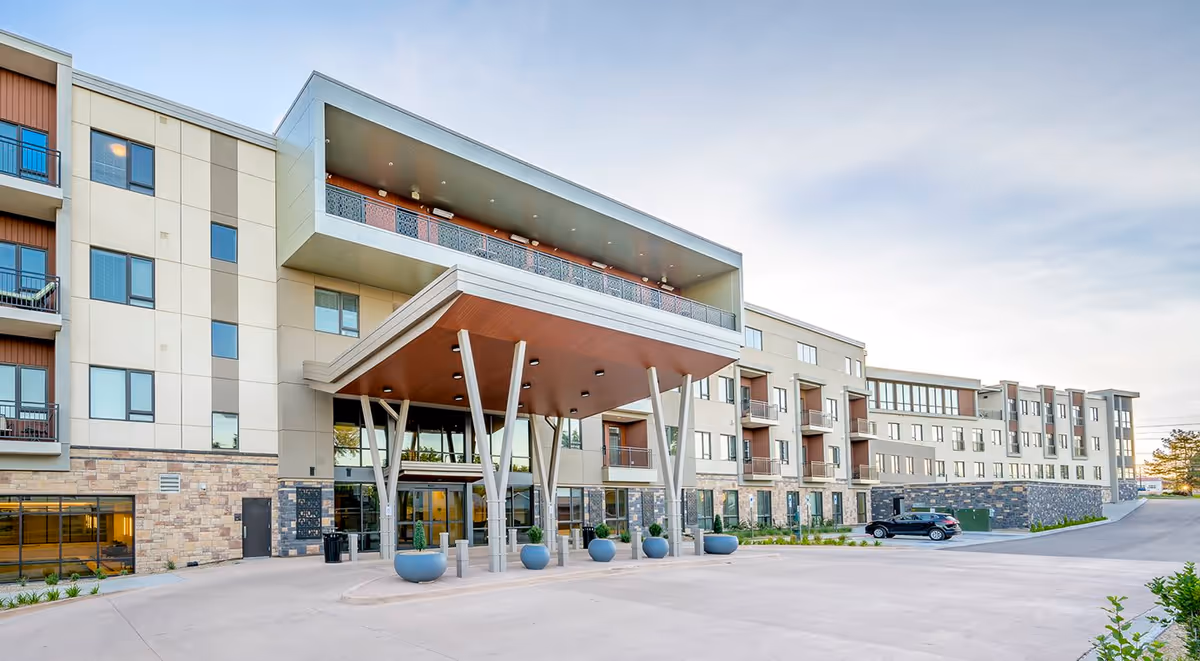 Front exterior view of a modern multi-story senior living facility with a covered entrance supported by angled pillars, large windows, balconies, and a parking area with a black car.