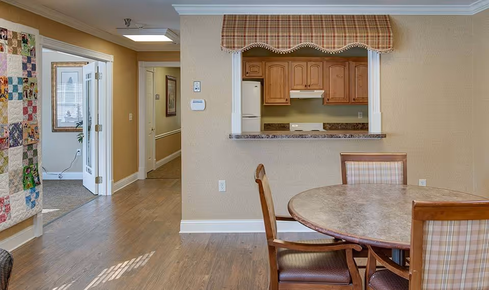 Interior dining area with a round table and chairs, a pass-through window to a small kitchen, and a hallway leading to other rooms.