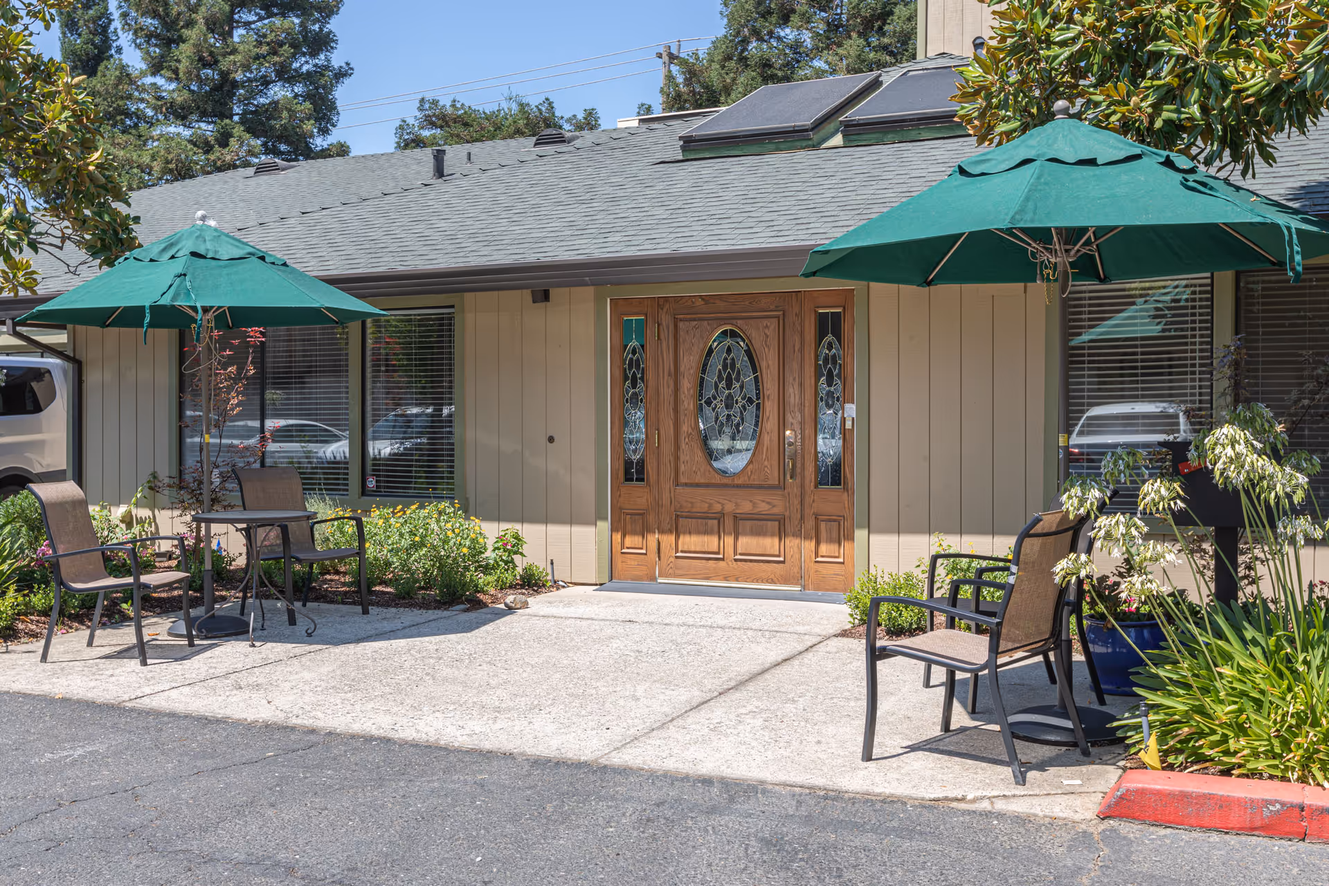 Front entrance of a single-story assisted living building with wooden double doors, patio tables, chairs, and green umbrellas.