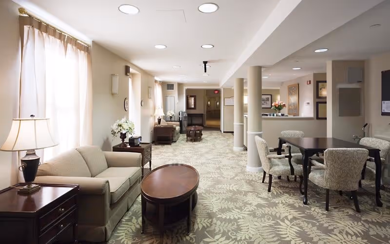 Spacious, well-lit communal lounge with sofas, armchairs, tables, lamps, and a reception desk down the hall.