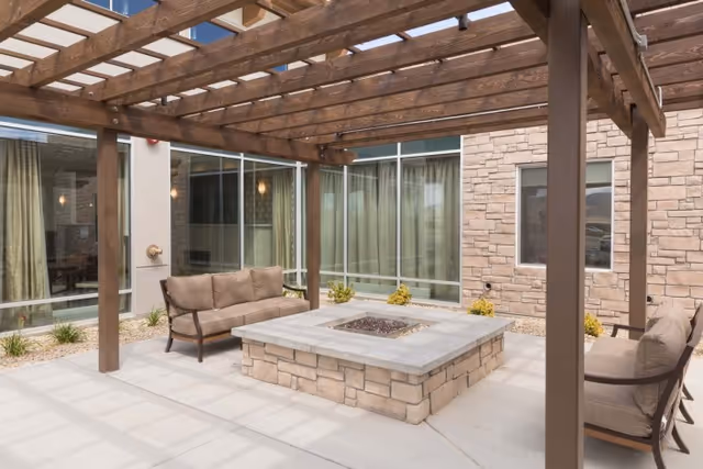 Outdoor patio area at Pointe Meadows with a wooden pergola overhead, cushioned seating around a square stone fire pit, and large windows with curtains on the building walls surrounding the patio.