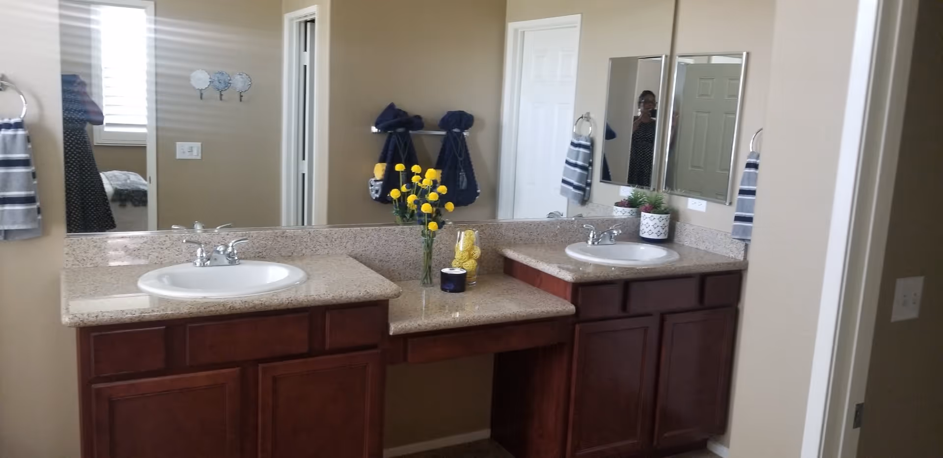 Double-sink bathroom vanity with granite countertops, mirrors, dark wood cabinets, towels, and small decorative plants and flowers.