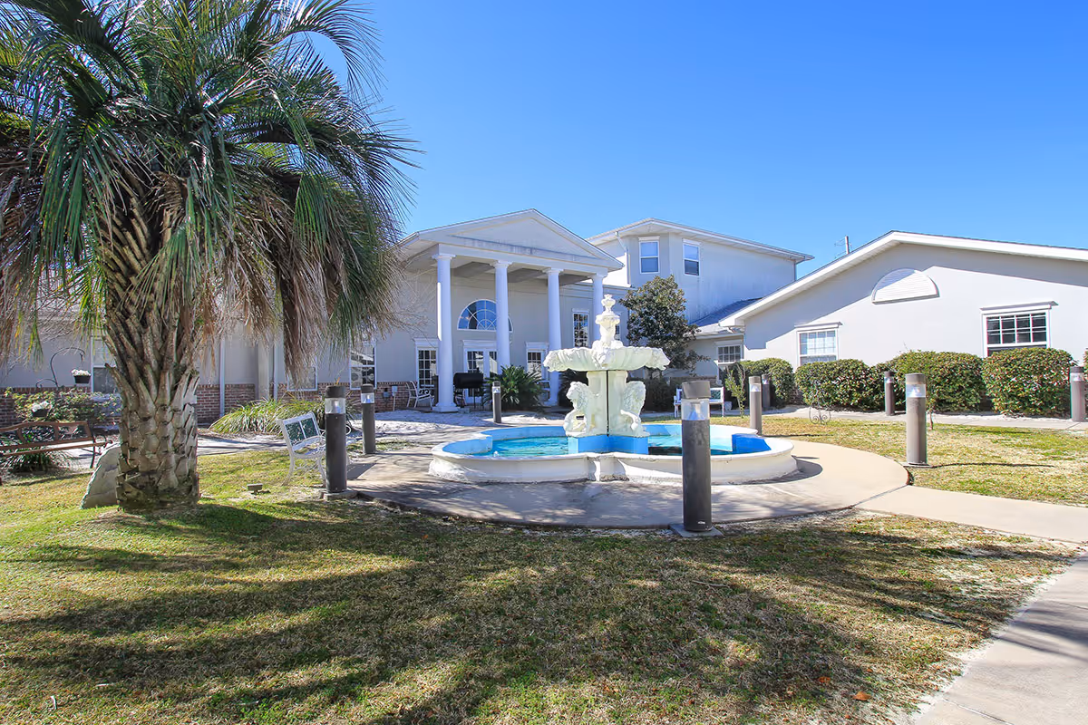 Outdoor courtyard area of Gulf Breeze Courtyard Assisted Living featuring a white decorative fountain in the center, surrounded by a circular walkway, green grass, palm trees, benches, and a white building with columns in the background under a clear blue sky.