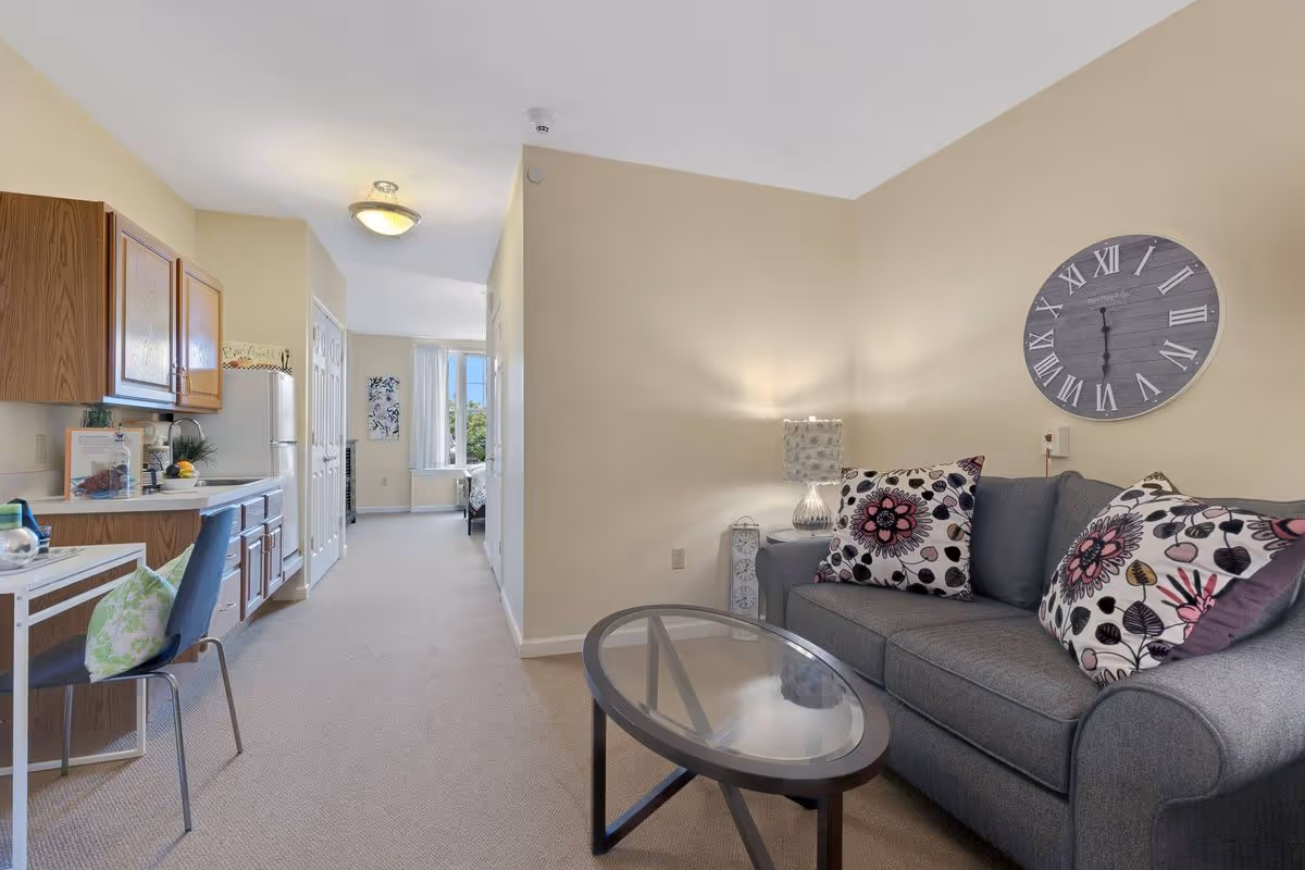 A cozy living area in a senior living facility featuring a gray sofa with floral patterned pillows, a glass coffee table, and a large wall clock with Roman numerals. To the left, there is a small dining area with a chair and a kitchen with wooden cabinets and a refrigerator. The space is well-lit with natural light coming from a window in the background.