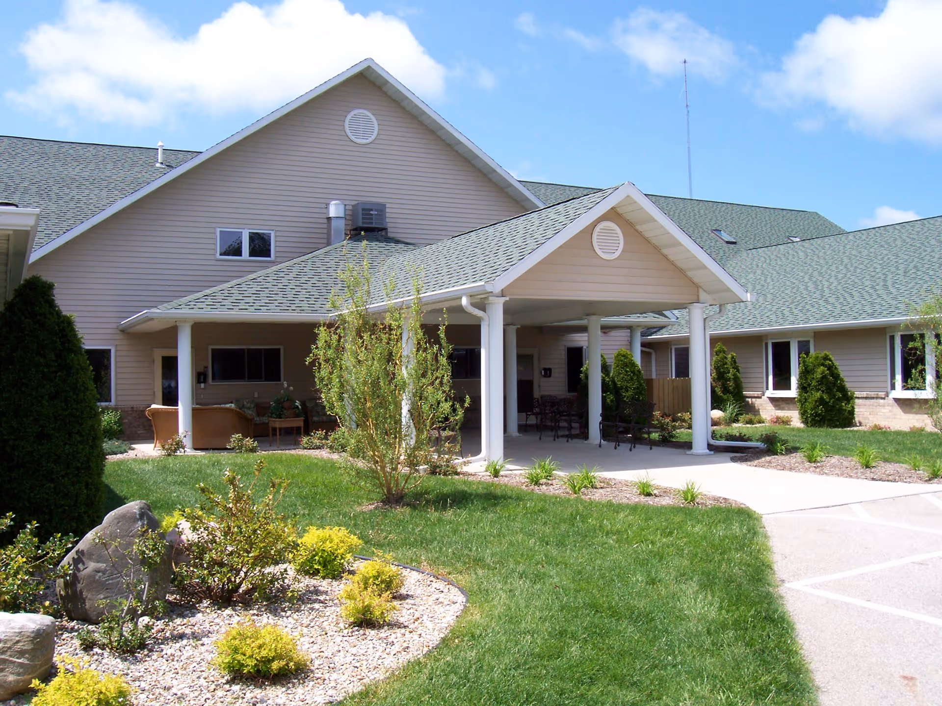 Front entrance of an assisted living building with a covered porte-cochere supported by white columns, outdoor seating, and landscaped lawn.