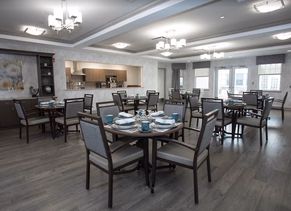 A spacious dining room with multiple round tables set with plates, napkins, glasses, and teal mugs. The room has wooden flooring, modern light fixtures on the ceiling, and large windows with curtains allowing natural light. In the background, there is a kitchen area visible through a large pass-through window.